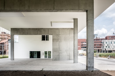 The image features a modern architectural structure with large concrete pillars and minimalistic design elements. In the background, several buildings with red brick and white geometric facades are visible. The foreground is dominated by the open space beneath the structure, which has a clean, urban aesthetic.