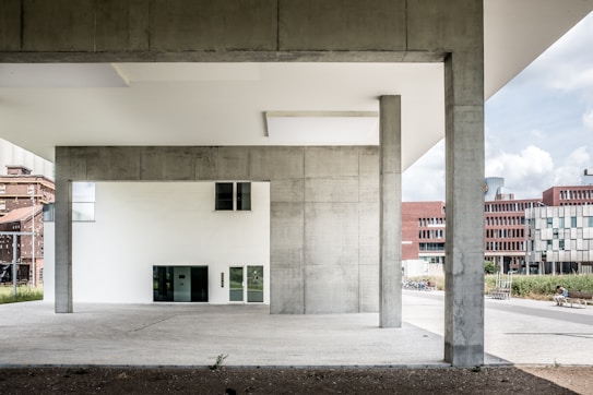 The image features a modern architectural structure with large concrete pillars and minimalistic design elements. In the background, several buildings with red brick and white geometric facades are visible. The foreground is dominated by the open space beneath the structure, which has a clean, urban aesthetic.