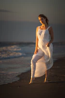 Elegant woman in a flowing white dress walking barefoot on a sandy beach at golden hour, soft waves in the background.