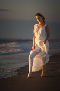 Model in a flowing dress standing barefoot on a sandy beach during golden hour.