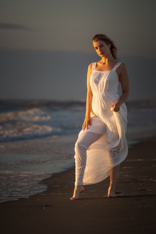 Model in a flowing dress standing barefoot on a sandy beach during golden hour.