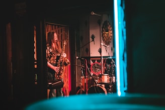 A musician with long hair plays the saxophone in a dimly lit room, surrounded by musical instruments like a drum set. The room has a vintage and eclectic decor with a prominent sunburst wall art and a warm color palette.