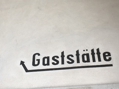 A beige wall features the word 'Gaststätte' painted in bold, black letters. An arrow points upward from the first letter, suggesting direction or movement.