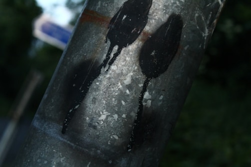 A close-up of a metal pole with dark paint streaks, resembling abstract figures against the worn surface. The background features a soft green blur, suggesting outdoor foliage or trees.