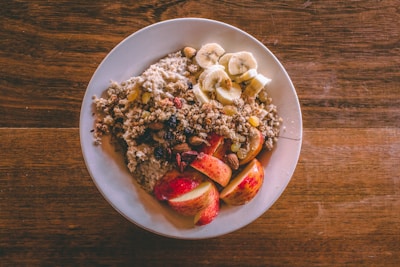 A white plate is filled with a healthy breakfast consisting of oatmeal topped with sliced bananas, chopped nuts, raisins, and apple slices. The oatmeal is garnished with granola, creating a texture-rich dish. The plate is set on a wooden table, adding a rustic feel to the presentation.