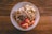 A white plate is filled with a healthy breakfast consisting of oatmeal topped with sliced bananas, chopped nuts, raisins, and apple slices. The oatmeal is garnished with granola, creating a texture-rich dish. The plate is set on a wooden table, adding a rustic feel to the presentation.