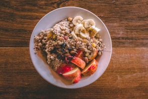 A white plate is filled with a healthy breakfast consisting of oatmeal topped with sliced bananas, chopped nuts, raisins, and apple slices. The oatmeal is garnished with granola, creating a texture-rich dish. The plate is set on a wooden table, adding a rustic feel to the presentation.