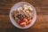 A white plate is filled with a healthy breakfast consisting of oatmeal topped with sliced bananas, chopped nuts, raisins, and apple slices. The oatmeal is garnished with granola, creating a texture-rich dish. The plate is set on a wooden table, adding a rustic feel to the presentation.
