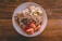 A white plate is filled with a healthy breakfast consisting of oatmeal topped with sliced bananas, chopped nuts, raisins, and apple slices. The oatmeal is garnished with granola, creating a texture-rich dish. The plate is set on a wooden table, adding a rustic feel to the presentation.