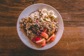 A white plate is filled with a healthy breakfast consisting of oatmeal topped with sliced bananas, chopped nuts, raisins, and apple slices. The oatmeal is garnished with granola, creating a texture-rich dish. The plate is set on a wooden table, adding a rustic feel to the presentation.