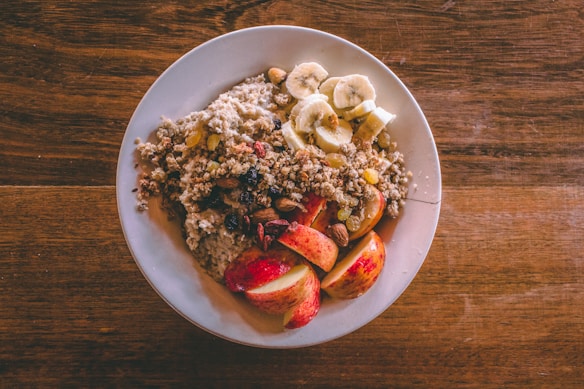 A white plate is filled with a healthy breakfast consisting of oatmeal topped with sliced bananas, chopped nuts, raisins, and apple slices. The oatmeal is garnished with granola, creating a texture-rich dish. The plate is set on a wooden table, adding a rustic feel to the presentation.