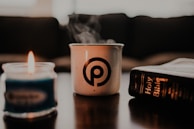 A steaming white mug with a circular logo, a blue candle with a flame, and a Holy Bible lie on a table with a blurred background.