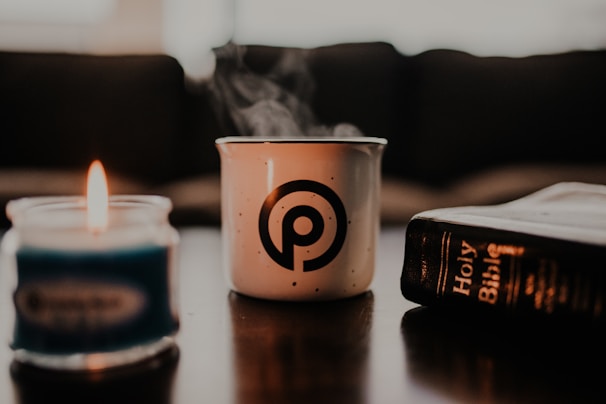 A steaming white mug with a circular logo, a blue candle with a flame, and a Holy Bible lie on a table with a blurred background.