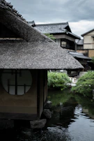 A traditional Japanese wooden house with a thatched roof stands by a calm pond. The architectural design features a round window, and the background includes more traditional structures and lush greenery. The sky is overcast, suggesting a serene and potentially rainy atmosphere.