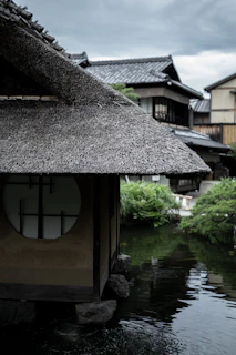 A traditional Japanese wooden house with a thatched roof stands by a calm pond. The architectural design features a round window, and the background includes more traditional structures and lush greenery. The sky is overcast, suggesting a serene and potentially rainy atmosphere.