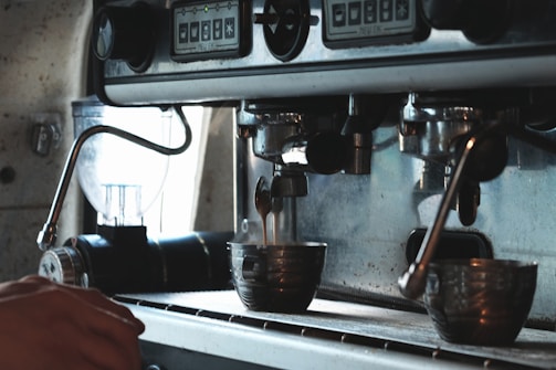 A close-up view of a coffee machine with two metallic cups positioned to receive freshly brewed coffee. The machine appears industrial with various levers and buttons on its front panel. A person's hand is visible on the left side of the image, suggesting interaction with the machine.