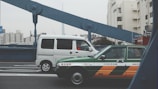 A green and orange taxi and a white van are driving over a bridge in an urban area. The bridge has distinctive large blue beams. In the background, there are several tall buildings, including a prominent tower.