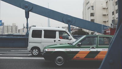A green and orange taxi and a white van are driving over a bridge in an urban area. The bridge has distinctive large blue beams. In the background, there are several tall buildings, including a prominent tower.