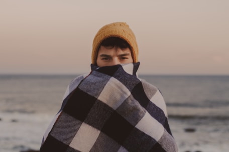 man covering his half-face with brown knit cap beside body of water at daytime