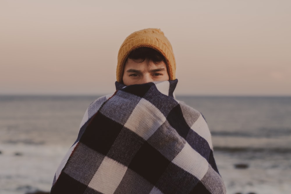 man covering his half-face with brown knit cap beside body of water at daytime