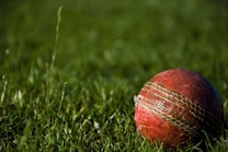 A well-used red cricket ball lies on a vibrant green grass field. The grass is lush, and the ball features prominent seam stitching. The image is slightly blurred in the background to enhance focus on the ball.