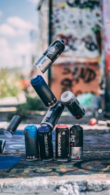 Several spray paint cans are creatively balanced on top of each other in an urban setting. The background is filled with colorful graffiti on walls, adding to the artistic atmosphere. Some cans are on the ground, and sunlight illuminates parts of the scene.