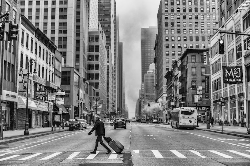 A busy urban street with tall skyscrapers lining both sides. A man is crossing the road, pulling a suitcase behind him. Several cars and buses are in motion, and street signs are visible. The image is in black and white, highlighting the architectural details and the bustling city atmosphere.