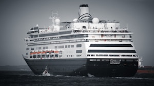 A large cruise ship is sailing on open water, featuring multiple decks and lifeboats along the side. The ship is primarily black and white with distinctive design elements and a name visible at the front.