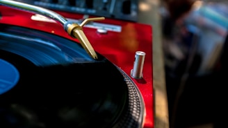 A close-up of a turntable with a vinyl record on it. The focus is on the stylus arm and the record is set against a background of a red surface with some audio equipment partially visible.