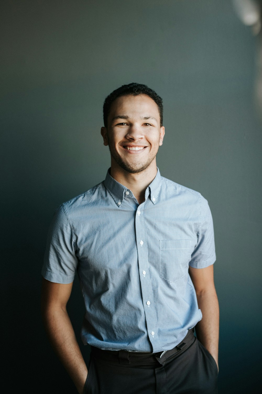 smiling man wearing blue and white gingham button-up collared shirt putting his hands on his pocket