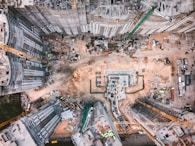 An aerial view of a large construction site surrounded by multiple high-rise buildings. Cranes are positioned around the site, with materials and machinery scattered across the ground. The area is bustling with activity, showcasing a mix of concrete, dirt, and metal.