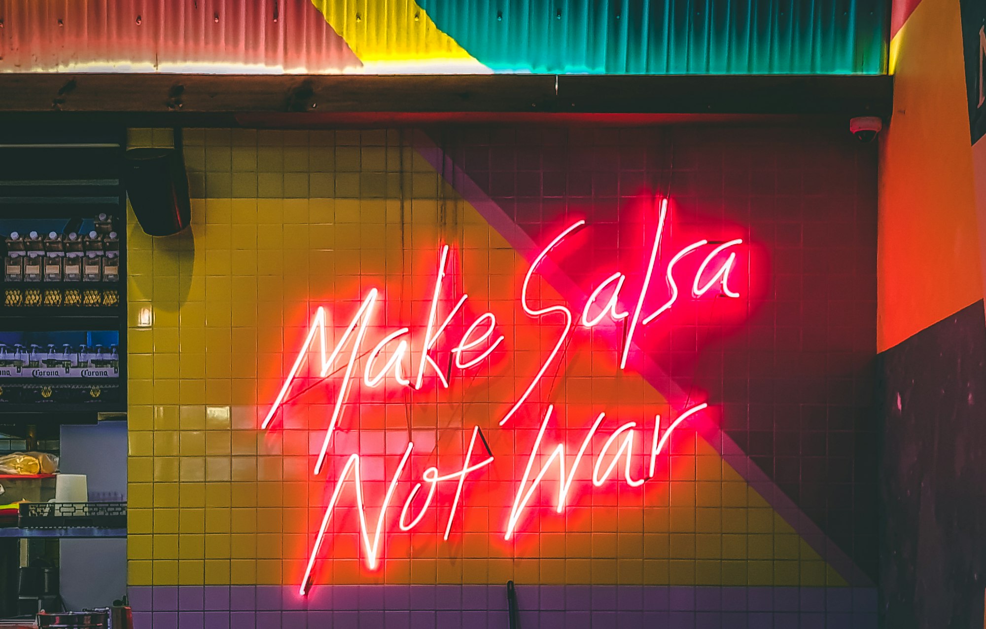 The words 'Make salsa not war' in a large, red neon sigh against a yellow tiled wall