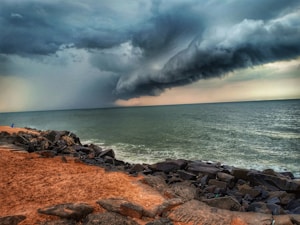 A dramatic seascape with ominous dark clouds forming a thick and swirling formation over the ocean, suggesting an approaching storm. The shoreline is lined with rocks and a sandy path, and the overall atmosphere is tense and turbulent.