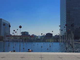 A calm urban scene featuring a pool of water with numerous tall, thin sculptures standing in it. These sculptures are metal poles with colorful circular disks on top. They are set against a backdrop of modern office buildings and an expansive blue sky. A person is sitting at the edge of the pool, seemingly relaxing or enjoying the view.