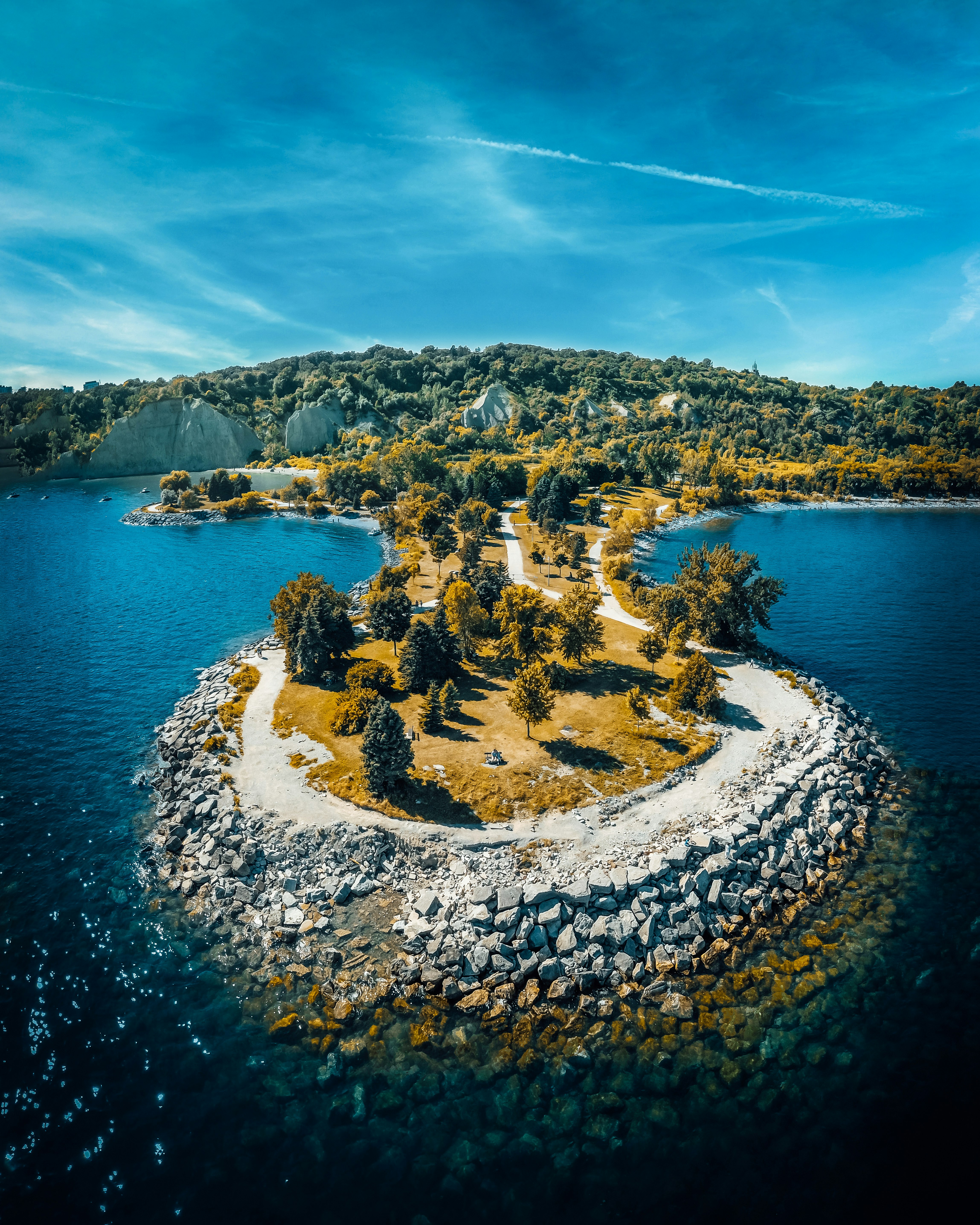 Aerial view of Bluffer's Park extending into Lake Ontario with paths winding through trees on the rocky breakwater, Scarborough Bluffs cliff faces visible in the background