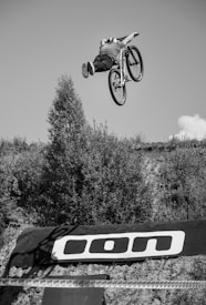 A cyclist is performing a high jump on a mountain bike above a dirt ramp, surrounded by trees and a clear sky. The rider is airborne, demonstrating a skillful trick, with their body stretched out behind the bike. A logo is visible on the ramp.