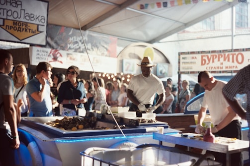 A bustling market scene with several people gathered around a seafood stall. Two men are preparing and selling seafood under a large canopy, surrounded by colorful stalls and banners. The atmosphere is lively with people wearing casual summer clothing, shopping, and interacting with vendors.