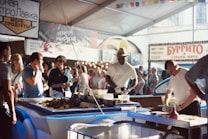 A bustling market scene with several people gathered around a seafood stall. Two men are preparing and selling seafood under a large canopy, surrounded by colorful stalls and banners. The atmosphere is lively with people wearing casual summer clothing, shopping, and interacting with vendors.