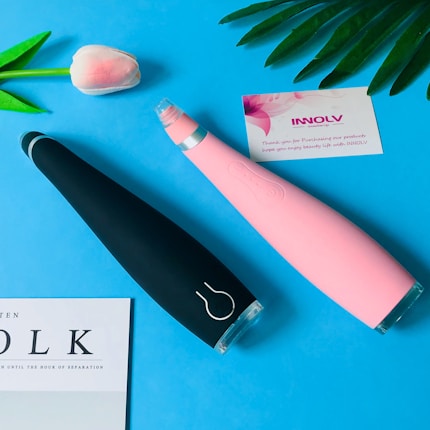 Two electronic beauty devices, one black and one pink, are placed diagonally on a blue surface. Nearby, a delicate pink and white artificial flower, a card with floral designs containing text, and a partially visible page with the word 'VOLK' are positioned.