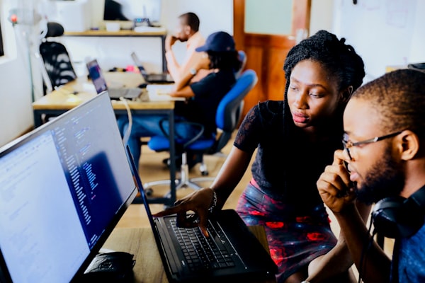 Two black people working on code together on a computer. In the background, two black people are working on their laptops.