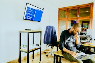A person wearing glasses and a hoodie is seated at a table, focused on typing on a laptop. The room has a modern and tech-oriented ambience with a screen mounted on the wall displaying lines of code. Shelves and cabinets are visible in the background, adding to the organized workspace setting.