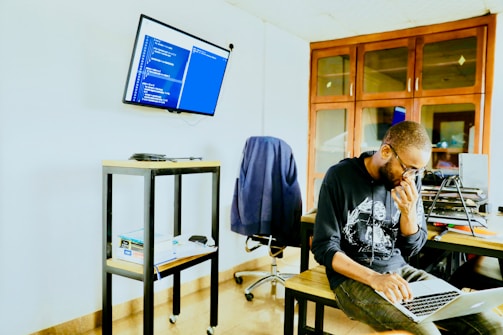 A person wearing glasses and a hoodie is seated at a table, focused on typing on a laptop. The room has a modern and tech-oriented ambience with a screen mounted on the wall displaying lines of code. Shelves and cabinets are visible in the background, adding to the organized workspace setting.