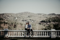 A couple sits on a white stone balustrade overlooking a lush, wooded landscape with rolling hills in the background. The setting is serene and romantic, with the man and woman holding each other closely, conveying intimacy and connection.