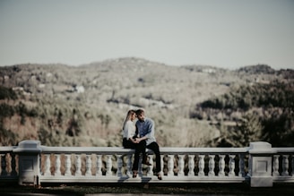 A couple sits on a white stone balustrade overlooking a lush, wooded landscape with rolling hills in the background. The setting is serene and romantic, with the man and woman holding each other closely, conveying intimacy and connection.