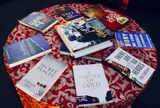 A round table covered with a red patterned cloth, displaying a variety of books with colorful covers. The books are arranged in a scattered manner, showcasing their covers and titles.