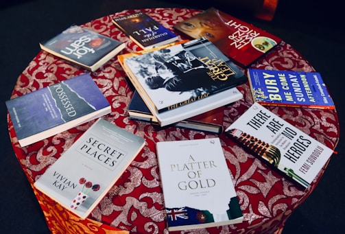 A round table covered with a red patterned cloth, displaying a variety of books with colorful covers. The books are arranged in a scattered manner, showcasing their covers and titles.