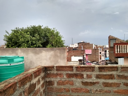 Brick buildings form a residential neighborhood under a cloudy sky. A prominent tree adds greenery, with a turquoise water tank visible on a roof.