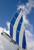 A sailing boat's mast and sail, prominently displaying large blue and white stripes, set against a clear sky with scattered clouds.