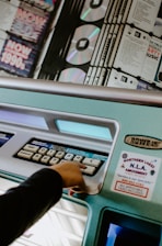 A hand is pressing a button on a jukebox, which features a display of CDs and various selection buttons labeled with numbers. The jukebox is teal in color and adorned with a sticker advertising an amusement service.