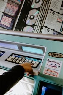 A hand is pressing a button on a jukebox, which features a display of CDs and various selection buttons labeled with numbers. The jukebox is teal in color and adorned with a sticker advertising an amusement service.